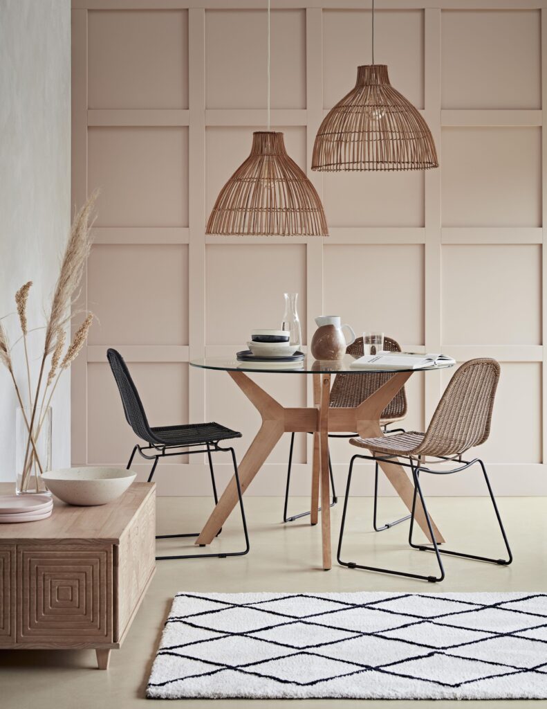 A styled dining space with a round glass top dining table, rattan chairs and pendant lights and soft wood tones, after the Japandi trend. Via: Habitat.