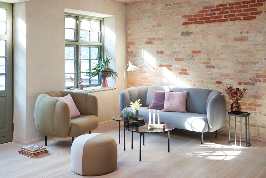 A living room with an exposed brick accent wall, Scandi design furniture in soft muted pastel colors that exude a Mediterranean aesthetic. Via Nest.co.uk.