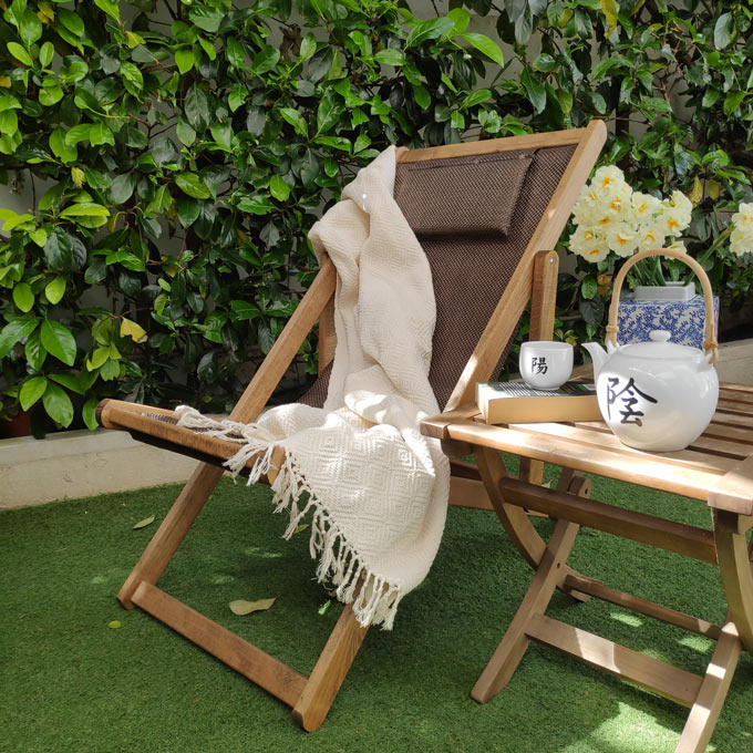 A wooden chaise longue styled with a throw next to a wooden side table in a green backyard.