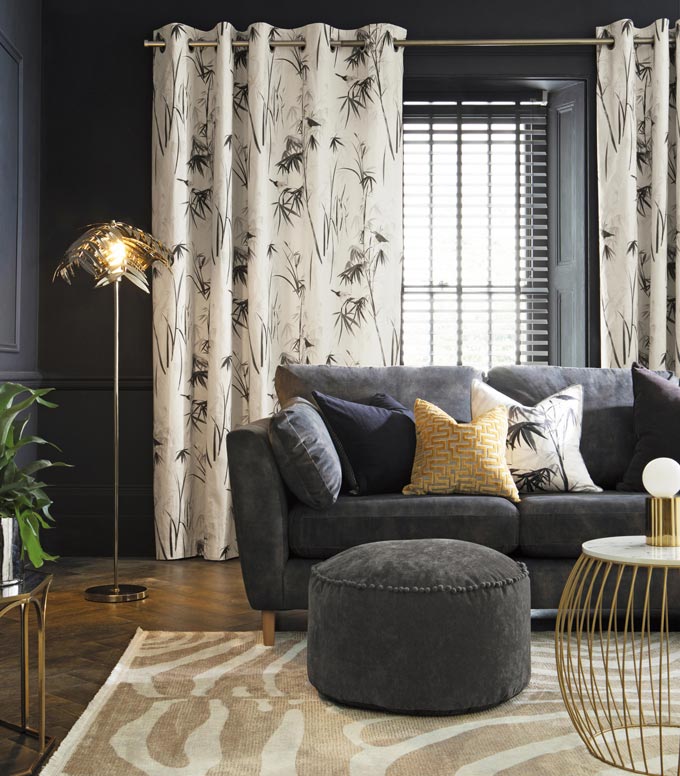 A dark, moody living room with a dark grey sofa and white pattern drapes in the background over a window door. Image via Next.