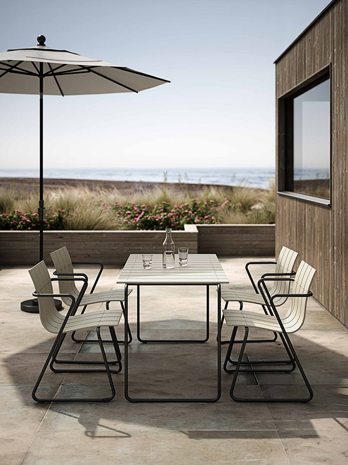 An outdoor dining set (table and chairs by Mater) in an off white hue, with a sun umbrella setup, overlooking at the sea in the background. Image: Mater.