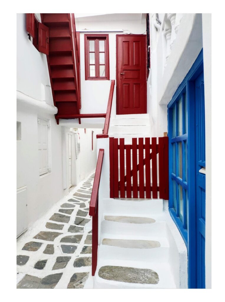 View of a narrow alley with white washed housing, red stairs and doorways and blue window doors.