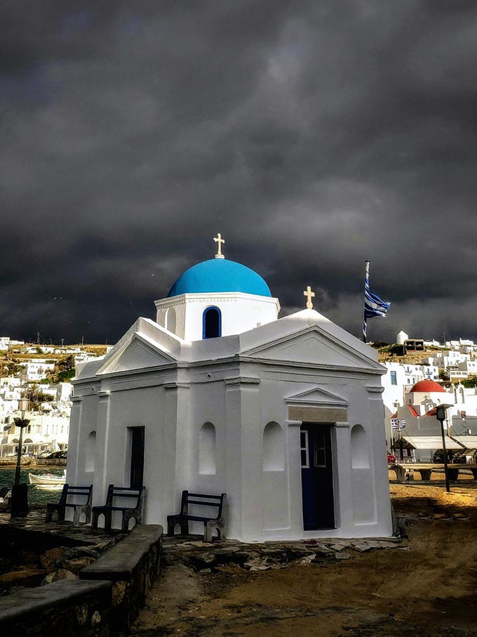 A white washed church with a blue dome in the foreground and a very black cloudy sky in the background.