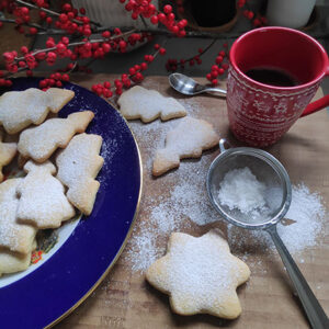 A pile of Velvet's Christmas Sugar Cookies, holly berries in the background and a red cup of hot chocolate next to the platter.