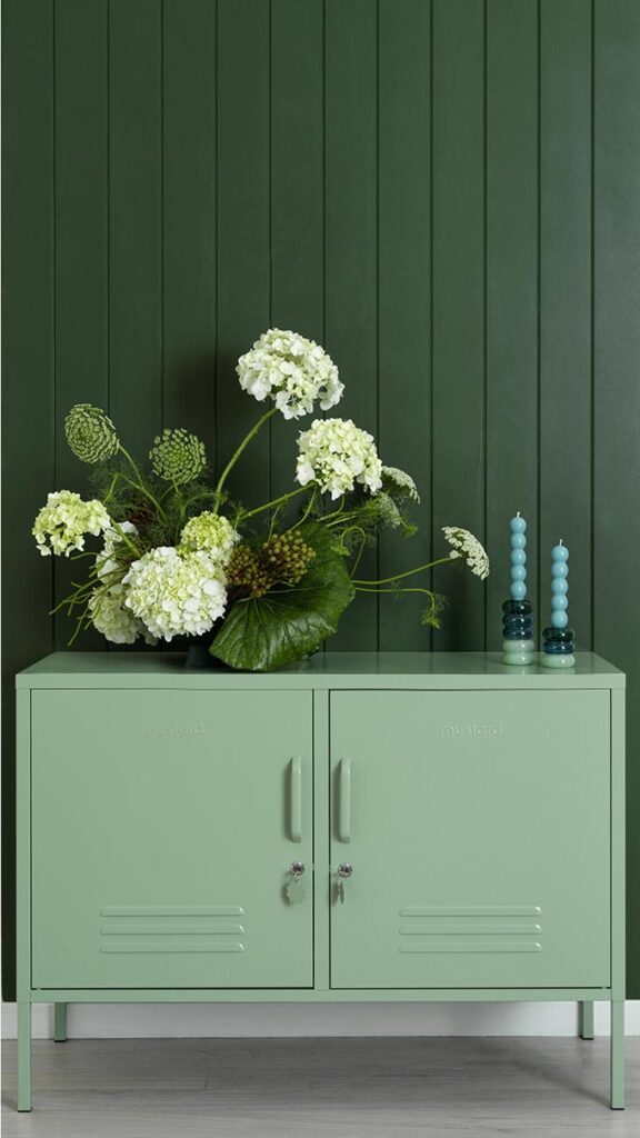 A sage green metal cabinet against dark green wood paneling with a flower arrangement atop. Image: Mustard.