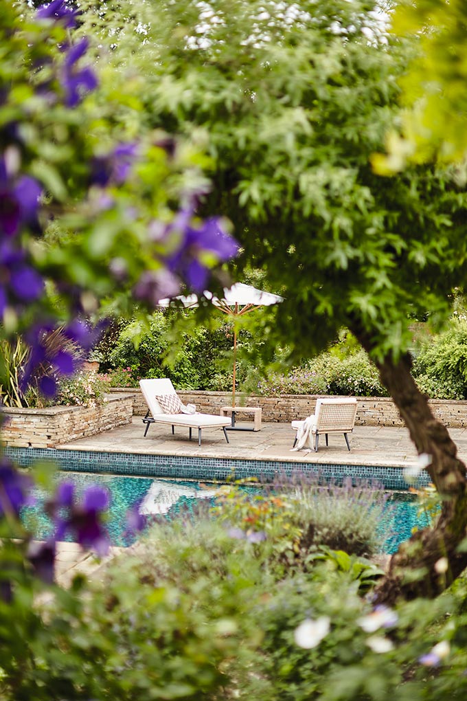 A peep hole view from a garden to a pool with two Cabrera sun loungers from OKA. Image: OKA UK.