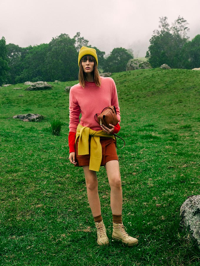 A lifestyle image of a woman dressed in sporty outfit somewhere outdoors with a long sleeve cashmere crew knit, shorts, hiking boots and hat. Imge: Oroton.