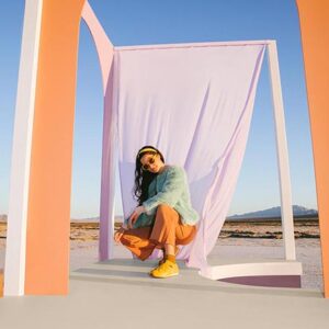 A lifestyle image of woman dressed in yellow Teva trainers, orange pants and cyan jacket. Image: TEVA.