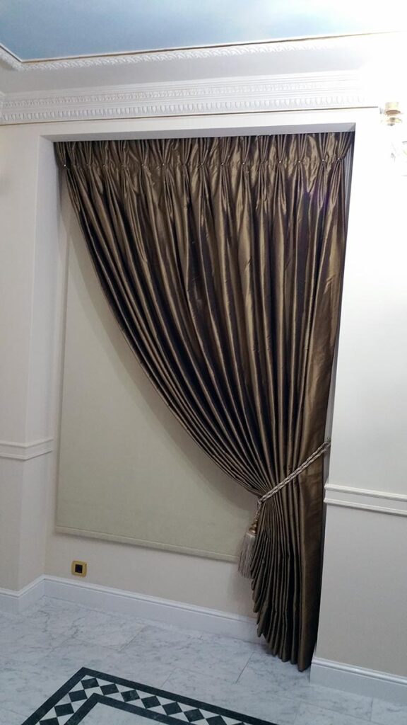 A formal dark satin pleated curtain with a tie back paired with a roller blind.
