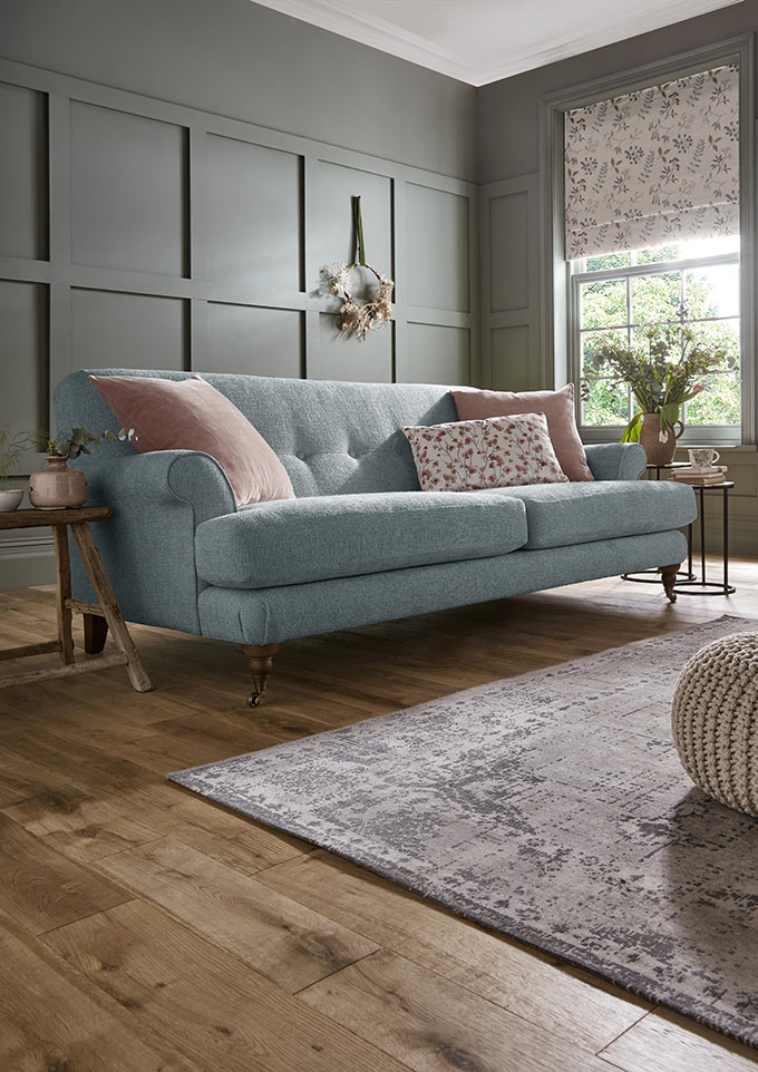 A beautiful light blue sofa with pink velvet throw pillows. What a relaxed space with all the light coming through the window that is covered by a Roman shade with a print. Image: Sofology.