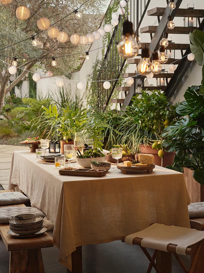 An outdoor garden dining setup with plenty of pots, planters, hanging lights and food on the table. Image: H&M Home.