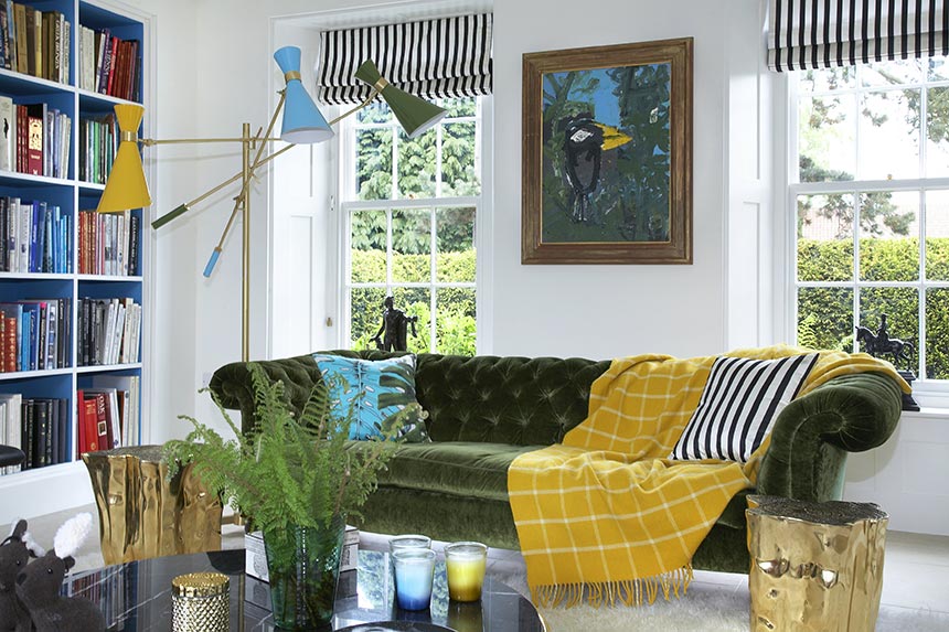 A vibrant and colorful living room with a velvet green sofa, a yellow throw in front of two windows with white an blue striped roman shades and a yellow, blue and green shade floor lamp. Image: Delightfull.