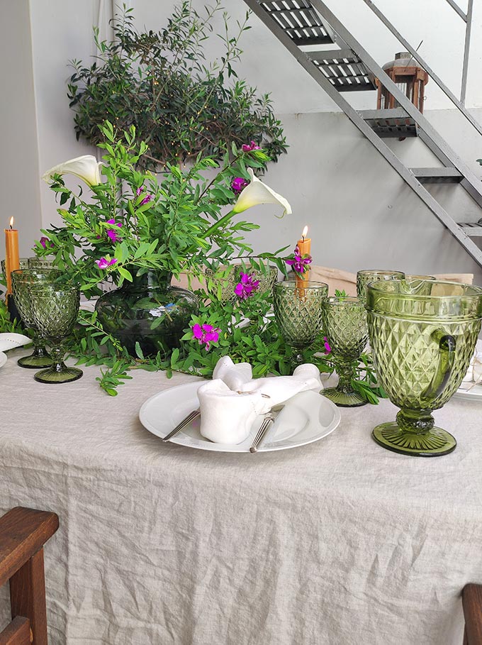 An outdoor dining setup for Easter lunch with lots of greenery as a centerpiece and a small olive tree in the background.