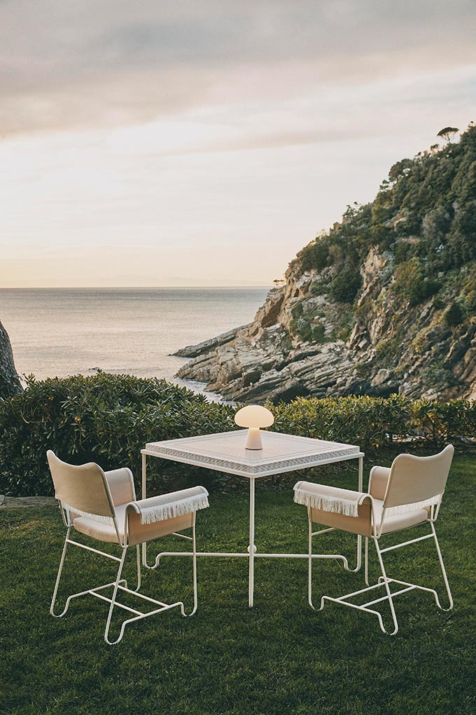 A lifestyle image of an outdoor setting with sea view featuring the Gubi Obello outdoor lamp on a square table. Image: Nest.co.uk.