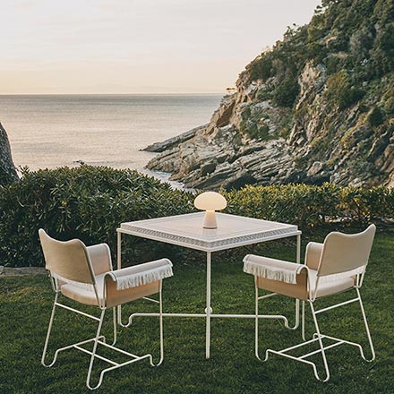 A lifestyle image of an outdoor setting with sea view featuring the Gubi Obello outdoor lamp on a square table. Image: Nest.co.uk.