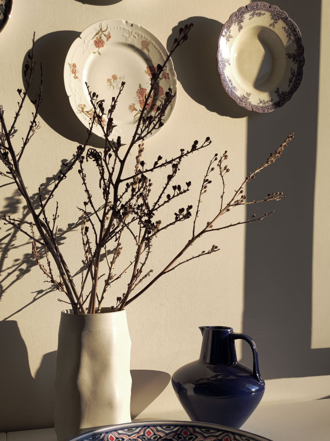 A ceramic white vase, a cobalt blue jug and a pattern platter resting on a white surface against the flooding natural light.