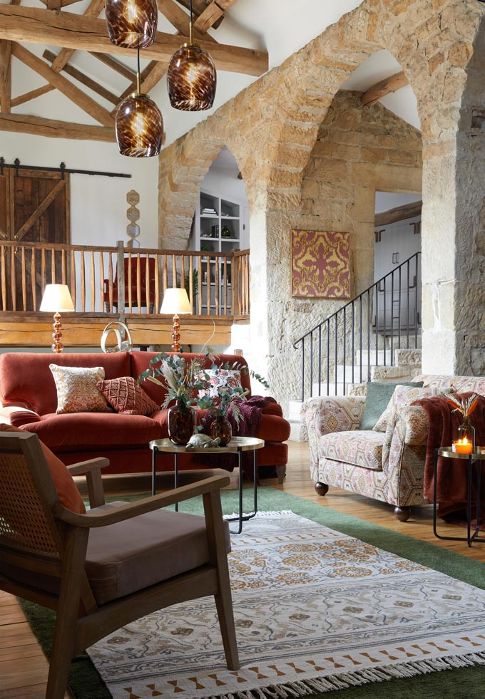 A stylish worldly sitting room with a deep peach velvet sofa, layered rugs, wooden architectural details and beautiful stone arched ways. Image: Dunelm.