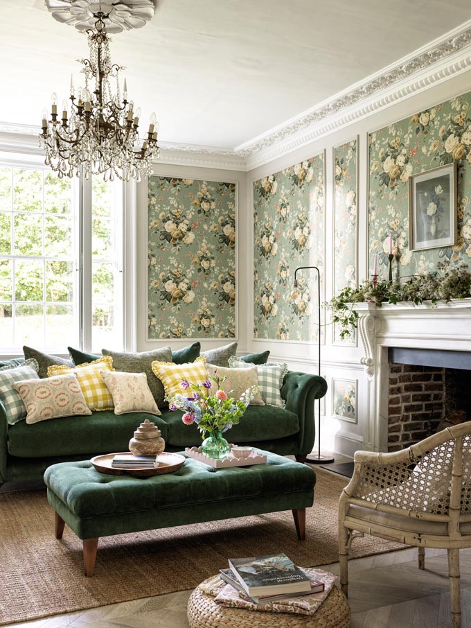 A Granny chic living room with a green velvet sofa, a fireplace, a hanging chandelier and flower pattern wallpaper. Image: Furniture Village.