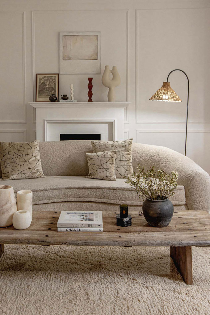 An off white boucle curvy sofa with a rustic coffee table, a beautiful Rattan Lamp by Katie Fischer and a fireplace with millwork in the background. Its warm and cozy, but it's not the Studio McGee signature look. Image via Katie Fischer GmbH.