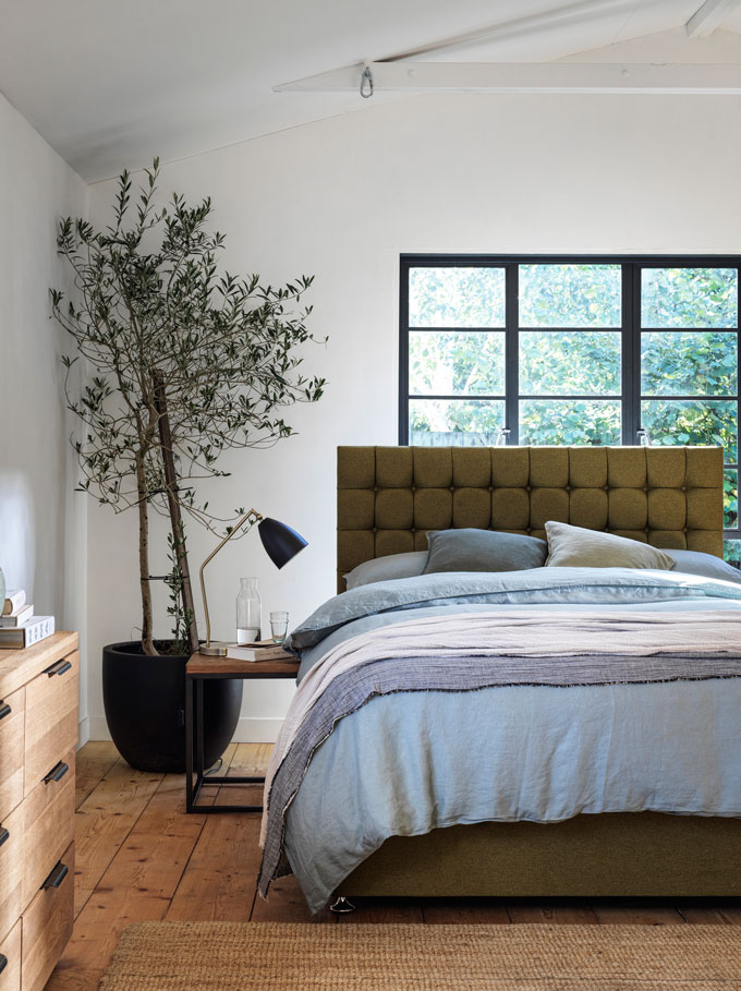 A modern version of a rustic bedroom with a white ceiling and white timber beams, an olive green bed with a cushioned headboard, wooden flooring and accessories and a large black planter in the corner. Image via Furniture Village.