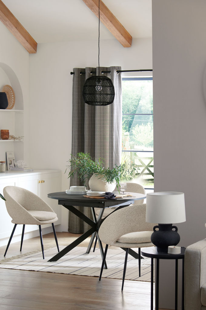 View of a bright, white dining space with a black round table, white chairs against a white background and exposed timber beams on the ceiling. Image via Next.