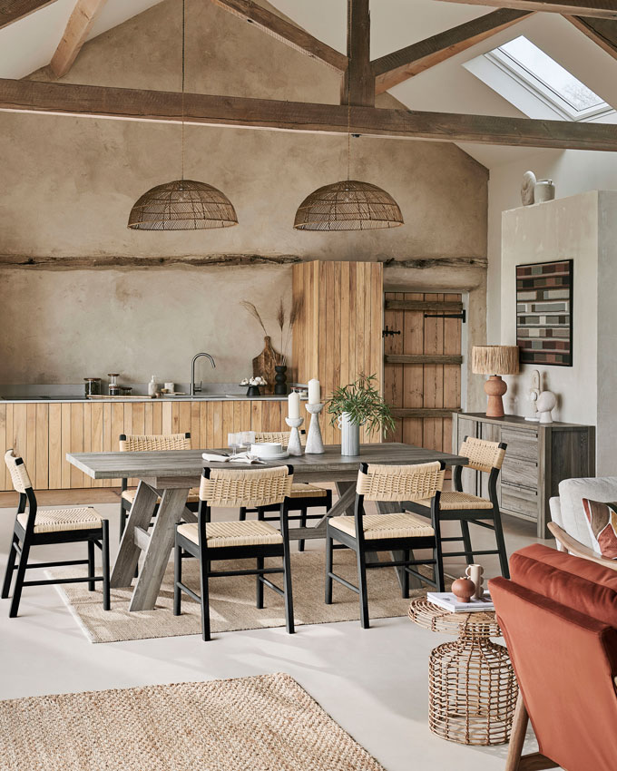 A contemporary kitchen with a rustic Farmhouse vibe, exposed timber beams in the pitched ceiling and a rustic dining table paired with black stained dining chairs. Image via Next.