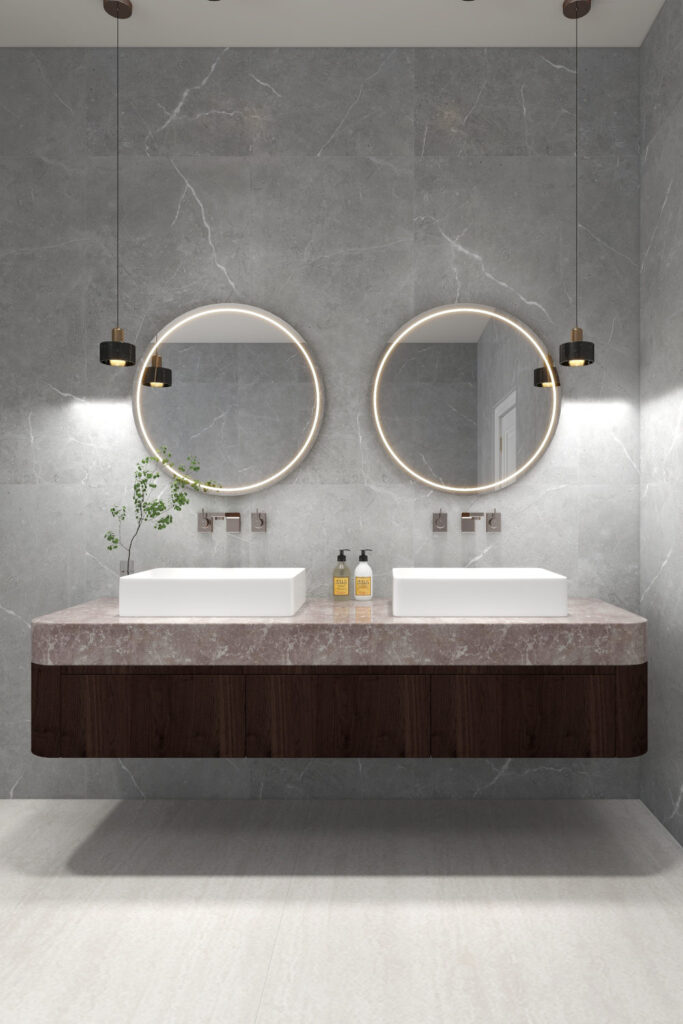 A contemporary bathroom with travertine flooring, a floating walnut vanity and a pink marble counter, double white over the counter basins, double mirrors, wall mounted faucets in rose gold and hanging pendants on left and right of the vanity. The background wall is a soft grey marble with white veins. Image copyright: Velvet Karatzas Interiors Atelier.